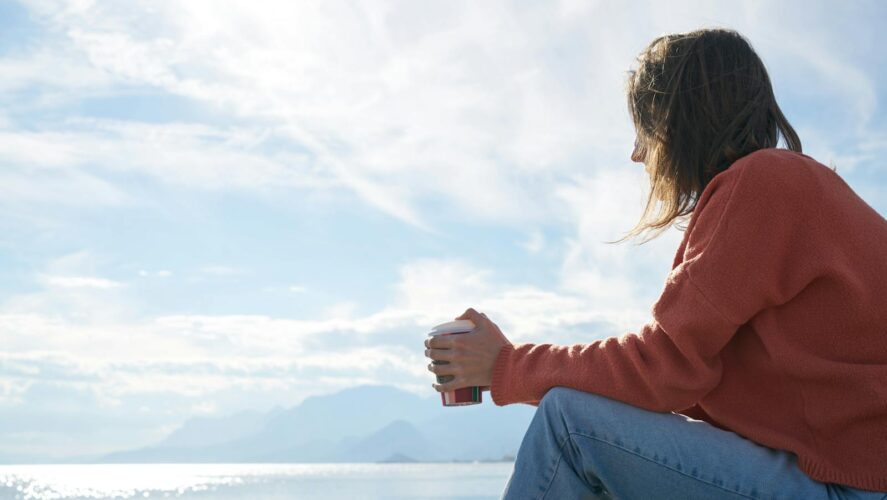 A woman in casual attire enjoys a hot drink while gazing at the sea, epitomizing relaxation.