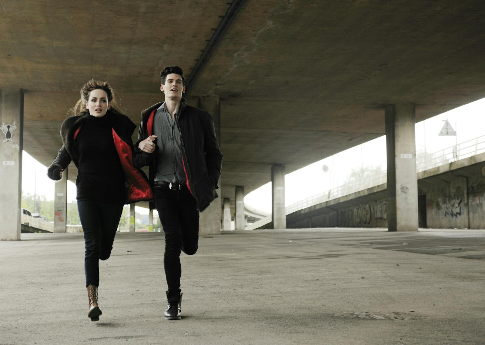 A couple runs energetically under a spacious urban bridge, capturing a sense of adventure and motion.