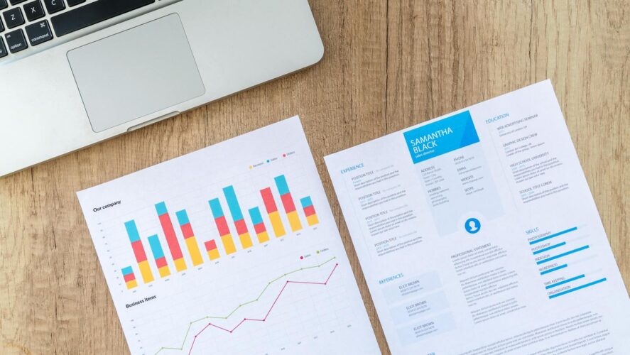Top view of a laptop, charts, and resume on a wooden desk, showcasing business analysis and job application.